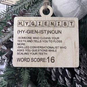 May include: A wooden Christmas ornament shaped like a Scrabble tile with the word "HYGIENIST" spelled out. The ornament also includes the definition of the word, "(HY-GIEN-IST)NOUN -SOMEONE WHO CLEANS YOUR TEETH AND TELLS YOU TO FLOSS MORE. -SKILLED CONVERSATIONALIST WHO ASKS YOU QUESTIONS WHILE SCALING YOUR TEETH." and the text "WORD SCORE:16".