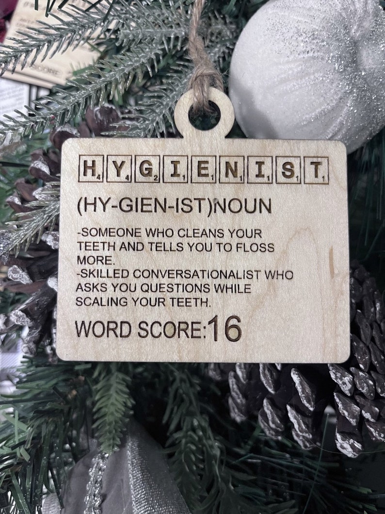May include: A wooden Christmas ornament shaped like a Scrabble tile with the word "HYGIENIST" spelled out. The ornament also includes the definition of the word, "(HY-GIEN-IST)NOUN -SOMEONE WHO CLEANS YOUR TEETH AND TELLS YOU TO FLOSS MORE. -SKILLED CONVERSATIONALIST WHO ASKS YOU QUESTIONS WHILE SCALING YOUR TEETH." and the text "WORD SCORE:16".