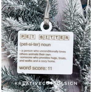 May include: White wooden ornament with the word "PET SITTER" spelled out in Scrabble tiles. The definition of a pet sitter is written below, along with the words "word score: 11". The bottom of the ornament reads "KREATIVECUTSDESIGN".