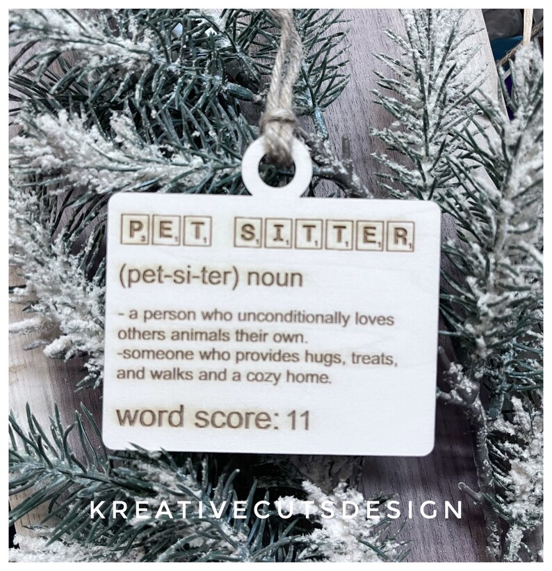 May include: White wooden ornament with the word "PET SITTER" spelled out in Scrabble tiles. The definition of a pet sitter is written below, along with the words "word score: 11". The bottom of the ornament reads "KREATIVECUTSDESIGN".