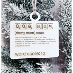 May include: Wooden ornament with the words "DOG MOM" in Scrabble tiles, with the definition "(dawg-mum) noun - has 99% of her camera roll of her dog. - someone willing to sacrifice their own freedoms to provide the lifestyle their dog expects and deserves. - provider of the best snacks, snuggles and unconditional love." and "word score: 12".