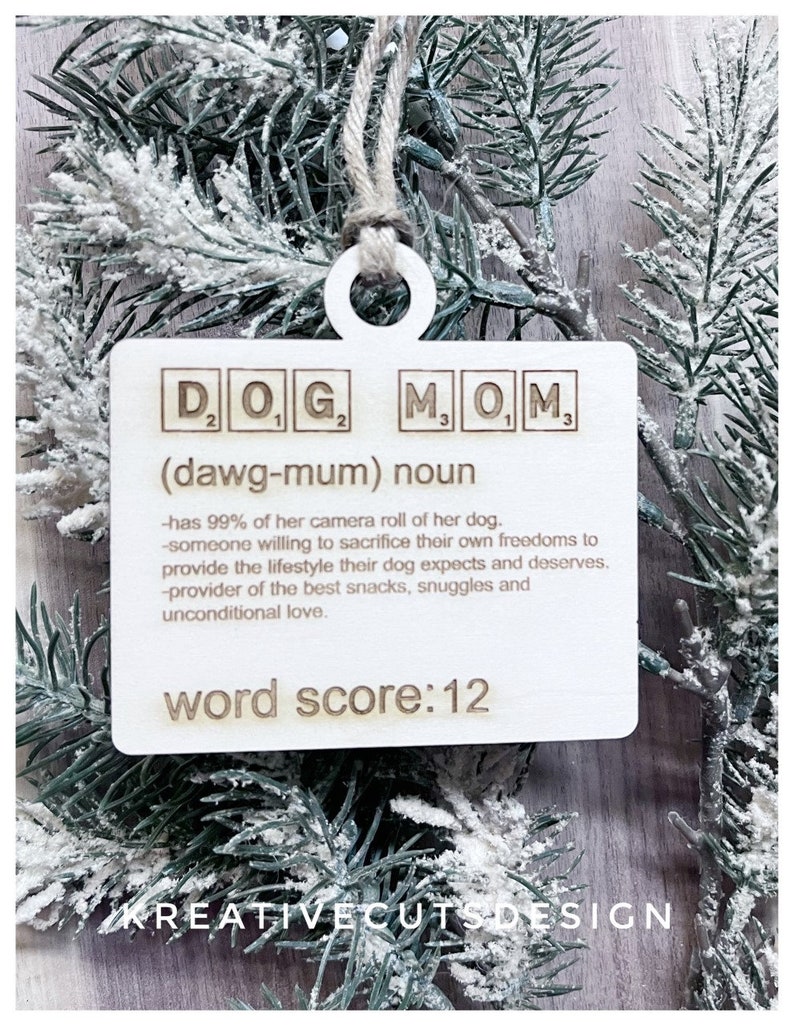 May include: Wooden ornament with the words "DOG MOM" in Scrabble tiles, with the definition "(dawg-mum) noun - has 99% of her camera roll of her dog. - someone willing to sacrifice their own freedoms to provide the lifestyle their dog expects and deserves. - provider of the best snacks, snuggles and unconditional love." and "word score: 12".