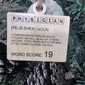 May include: Wooden ornament with the word "PHYSICIAN" spelled out in Scrabble tiles. The ornament also includes the definition of the word, "(FE-ZI-SHEN) NOUN -COMPASSIONATE PERSON WHO IS FOCUSED ON WELL-BEING OF OTHERS. -NATURAL BORN PROBLEM SOLVER WHO OFTEN THINKS OUTSIDE THE BOX." and a word score of 19.