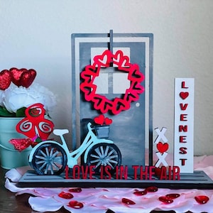 May include: A wooden Valentine's Day decoration featuring a red heart wreath, a bicycle with a basket of red hearts, and the text "LOVE IS IN THE AIR".