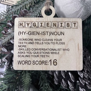 May include: A wooden Christmas ornament shaped like a Scrabble tile with the word "HYGIENIST" spelled out. The ornament also includes the definition of the word, "(HY-GIEN-IST)NOUN -SOMEONE WHO CLEANS YOUR TEETH AND TELLS YOU TO FLOSS MORE. -SKILLED CONVERSATIONALIST WHO ASKS YOU QUESTIONS WHILE SCALING YOUR TEETH." and the text "WORD SCORE:16".