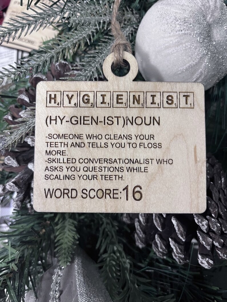 May include: A wooden Christmas ornament shaped like a Scrabble tile with the word "HYGIENIST" spelled out. The ornament also includes the definition of the word, "(HY-GIEN-IST)NOUN -SOMEONE WHO CLEANS YOUR TEETH AND TELLS YOU TO FLOSS MORE. -SKILLED CONVERSATIONALIST WHO ASKS YOU QUESTIONS WHILE SCALING YOUR TEETH." and the text "WORD SCORE:16".