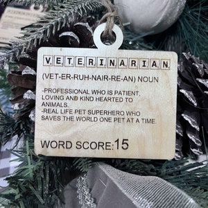 May include: Wooden ornament shaped like a Scrabble tile with the word "VETERINARIAN" spelled out. The ornament also includes the definition of the word, "(VET-ER-RUH-NAIR-RE-AN) NOUN -PROFESSIONAL WHO IS PATIENT, LOVING AND KIND HEARTED TO ANIMALS. -REAL LIFE PET SUPERHERO WHO SAVES THE WORLD ONE PET AT A TIME." The ornament also includes the text "WORD SCORE: 15".