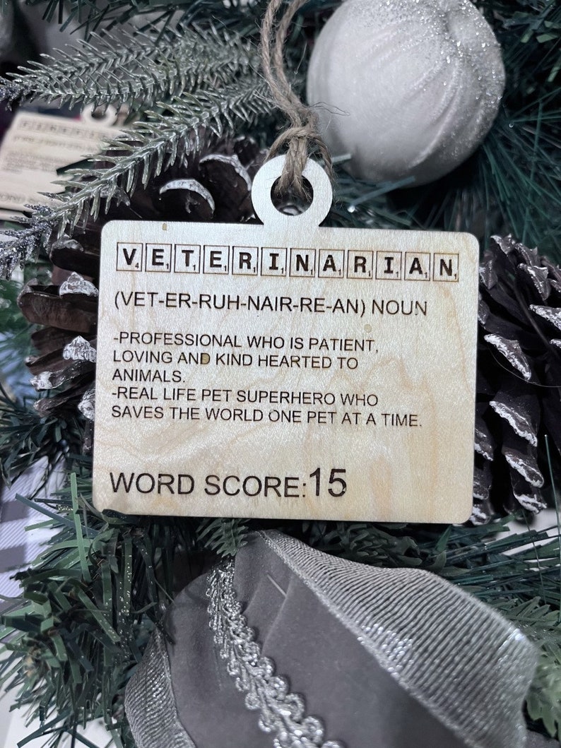 May include: Wooden ornament shaped like a Scrabble tile with the word "VETERINARIAN" spelled out. The ornament also includes the definition of the word, "(VET-ER-RUH-NAIR-RE-AN) NOUN -PROFESSIONAL WHO IS PATIENT, LOVING AND KIND HEARTED TO ANIMALS. -REAL LIFE PET SUPERHERO WHO SAVES THE WORLD ONE PET AT A TIME." The ornament also includes the text "WORD SCORE: 15".