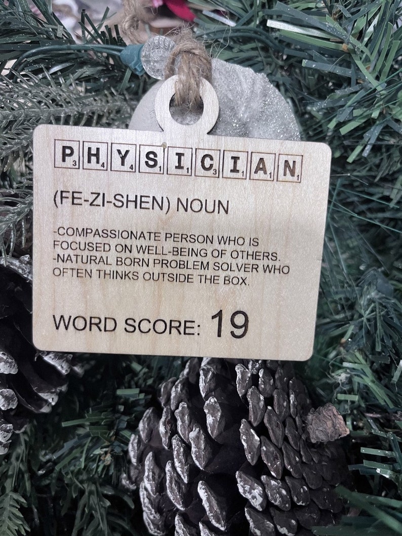May include: A wooden ornament shaped like a tag with the word "PHYSICIAN" spelled out in Scrabble tiles. The tag also includes the definition of the word, "(FE-ZI-SHEN) NOUN -COMPASSIONATE PERSON WHO IS FOCUSED ON WELL-BEING OF OTHERS. -NATURAL BORN PROBLEM SOLVER WHO OFTEN THINKS OUTSIDE THE BOX." and the text "WORD SCORE: 19".