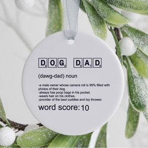 May include: White ceramic ornament with the word "DOG" and "DAD" spelled out in Scrabble tiles. The definition of "dawg-dad" is written below the tiles. The definition reads: "a male owner whose camera roll is 99% filled with photos of their dog. -always has poop bags in his pocket. -wears hair on his clothes. -provider of the best cuddles and toy thrower." The ornament also includes the text "word score: 10".