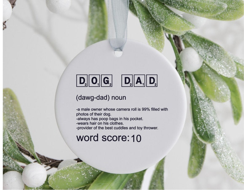 May include: White ceramic ornament with the word "DOG" and "DAD" spelled out in Scrabble tiles. The definition of "dawg-dad" is written below the tiles. The definition reads: "a male owner whose camera roll is 99% filled with photos of their dog. -always has poop bags in his pocket. -wears hair on his clothes. -provider of the best cuddles and toy thrower." The ornament also includes the text "word score: 10".