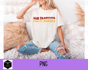 Puede incluir: Una camiseta blanca con el texto "SAN FRANCISCO FORTY NINERS" en letras rojas y doradas. La camiseta la lleva puesta una persona sentada en una superficie peluda blanca y marrón.