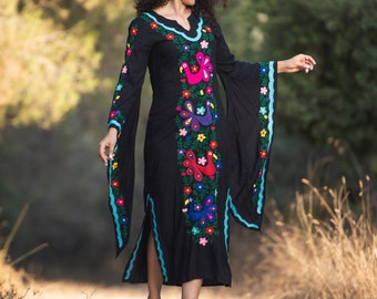 May include: A woman wearing a black maxi dress with colorful embroidered flowers and birds. The dress has long sleeves and a flowing silhouette.