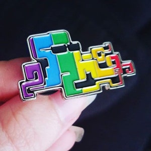 May include: A rainbow colored enamel pin shaped like a pixelated character. The pin is made of metal and has a silver finish.