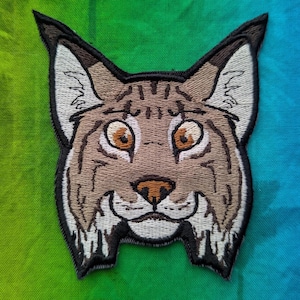 Puede incluir: Un parche bordado con la cara de un lince. El lince tiene ojos marrones y un hocico blanco.
