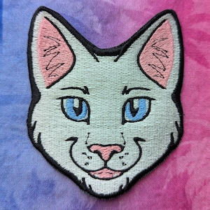 May include: A white cat with blue eyes and pink ears is embroidered on a light blue and pink tie-dye background.