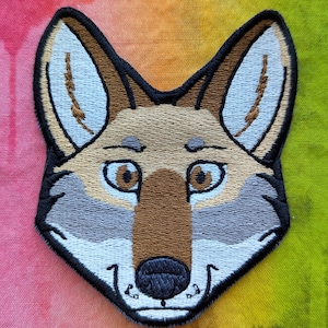May include: Embroidered patch of a coyote's face with brown fur, a grey muzzle, and black nose. The patch is on a colourful tie-dye background.