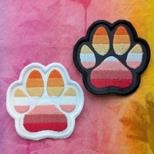 Puede incluir: Dos parches bordados con huellas de patas. Uno tiene un borde blanco con rayas naranjas, amarillas, rosas y rojas. El otro tiene un borde negro con rayas naranjas, amarillas, rosas y rojas. El fondo es un degradado de rosa, morado y verde.
