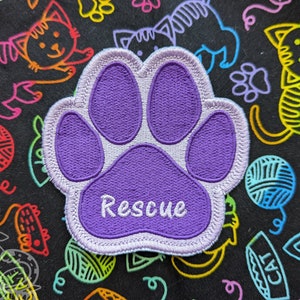 Puede incluir: Parche bordado con forma de huella de pata de color morado con costuras blancas. El parche tiene la palabra "Rescue" escrita en blanco en la parte inferior.
