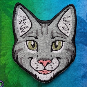 May include: Embroidered patch of a grey cat's face with green eyes, a pink nose, and a smiling mouth. The patch has a black border and is set against a green and blue gradient background with palm tree silhouettes.