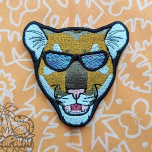 May include: Embroidered patch featuring a cougar wearing sunglasses. The patch has a black border, with the cougar's face in shades of brown, tan, and blue. The sunglasses are a vibrant blue and purple. The patch is on an orange background with a white floral pattern.
