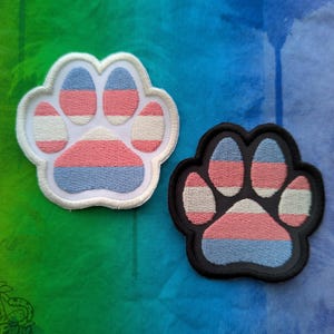 May include: Two embroidered paw print patches. One has a white border, the other a black border. Both feature a design in the colors of the transgender pride flag: pink, white, and blue. The patches are likely for clothing or accessories.