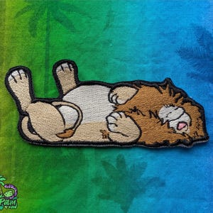 May include: Embroidered patch featuring a cartoon lion lying on its back with its paws in the air. The lion has a brown mane, tan paws, and a white belly. The patch has a black border and is on a green and blue background.