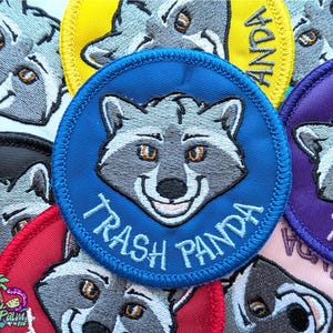 Puede incluir: Parches bordados con un diseño de mapache sonriente. El parche principal es azul con la inscripción "Trash Panda". Otros parches son amarillos, morados, rojos y rosas, todos con la misma cara de mapache.