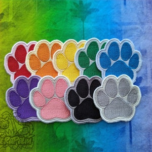 Puede incluir: Nueve parches bordados con forma de huella de pata en varios colores, incluyendo rojo, naranja, amarillo, verde, azul, morado, rosa, negro y gris. Los parches están dispuestos en un semicírculo.