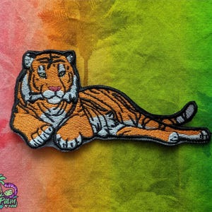 Puede incluir: Un parche bordado que muestra un tigre tumbado en posición de descanso. El tigre tiene pelaje naranja con rayas negras, detalles blancos en la cara y las patas, y una nariz rosa. El parche está delineado con hilo negro y tiene textura.