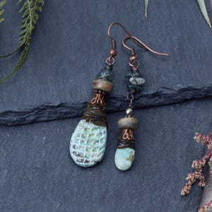Op de afbeelding: Handgemaakte ambachtelijke oorbellen met een uniek ontwerp. De oorbellen hebben een constructie van koperdraad en kralen, met turquoise en groene stenen accenten. De grotere oorbel heeft een getextureerde, druppelvormige turkooizen steen.