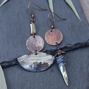 Op de afbeelding: Handgemaakte koperen oorbellen met een uniek ontwerp. Elke oorbel heeft een gehamerd koperen element, een beschilderd keramisch stuk met een landschapsscène en een decoratieve hanger met blauwe stof en een puntige, iriserende kraal.