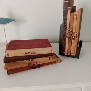 May include: A set of wooden cutting boards with engraved text, including "Julienne," "Dice," and "Chop." The boards vary in color, with a mix of light and dark wood tones. A black metal stand holds a collection of wooden blocks with similar text.