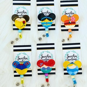 Puede incluir: Seis portagajes retráctiles con temática de princesas Disney con detalles de purpurina. Cada portagajes presenta una princesa diferente con un diseño de orejas de Mickey Mouse. Las princesas son Bella, Jasmine, Moana, Blancanieves, Ariel y Cenicienta. Los portagajes están unidos a tarjetas con rayas blancas y negras con el texto "Smashin Creations" en ellas.