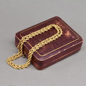 May include: A gold-colored rope chain necklace rests on a vintage maroon jewelry box. The necklace is made of tightly woven links, creating a textured appearance. The box has a rectangular shape with a slightly worn, aged look.