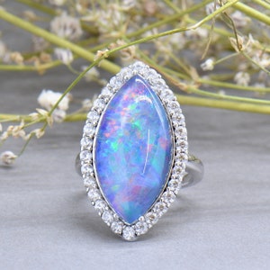 May include: A silver ring with a large, iridescent opal set in a marquise shape. The opal is surrounded by a halo of smaller, clear stones.
