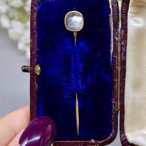 May include: A vintage gold-toned stick pin with a square, iridescent, moonstone-like cabochon. The pin is displayed in a dark blue velvet-lined box with a burgundy exterior. The box is open, revealing the pin's delicate design.
