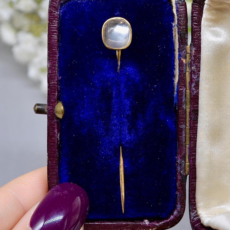 May include: A vintage gold-toned stick pin with a square, iridescent, moonstone-like cabochon. The pin is displayed in a dark blue velvet-lined box with a burgundy exterior. The box is open, revealing the pin's delicate design.