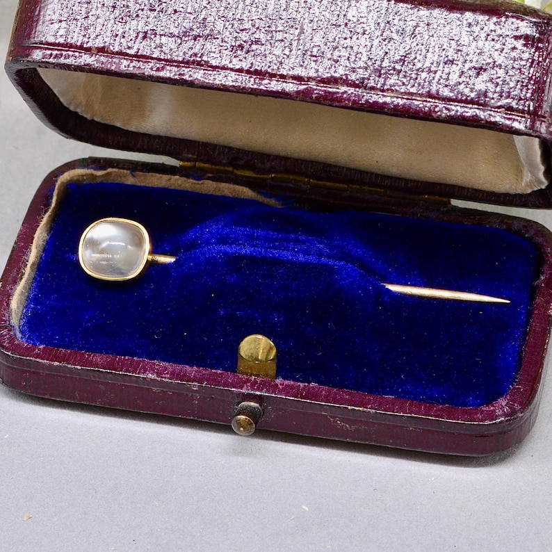 May include: A vintage gold-tone stick pin with a square, iridescent, pearl-like stone. The pin is displayed in an open, burgundy leatherette box with a blue velvet interior. The box is closed with a gold-tone clasp.