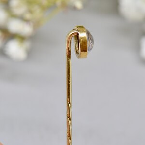 May include: A vintage gold-tone stick pin with a clear, round gemstone set in a gold bezel. The pin has a twisted design on the lower portion and a curved top. The background is blurred, with soft white and green tones.