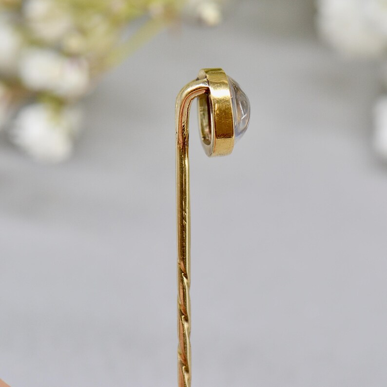 May include: A vintage gold-tone stick pin with a clear, round gemstone set in a gold bezel. The pin has a twisted design on the lower portion and a curved top. The background is blurred, with soft white and green tones.