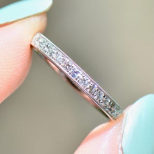 Può includere: Primo piano di un delicato anello argentato. L'anello è adornato da una fila di piccole pietre preziose scintillanti. La fascia presenta un design dettagliato e strutturato. L'anello è tenuto tra due dita.