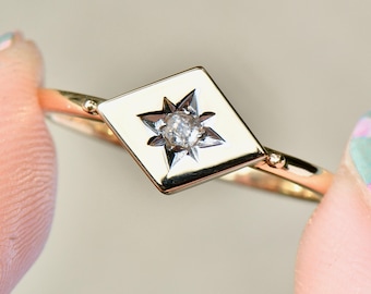 May include: A gold ring featuring a diamond-shaped setting with a central diamond and a starburst design. The ring's band is thin and gold, with small decorative details. The ring is held between two fingers.