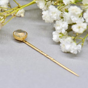 May include: A gold-colored hair pin with a clear, oval-shaped stone at the top. The pin has a textured design on the shaft. White flowers are in the background, resting on a gray surface. This accessory is suitable for various hairstyles.
