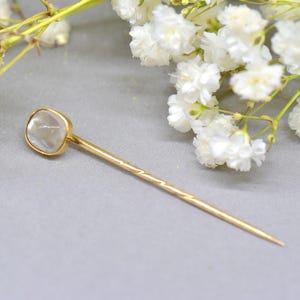 May include: A gold-colored hat pin with a square, iridescent stone at the top. The pin has a textured design on the shaft and a pointed end. White flowers are in the background.