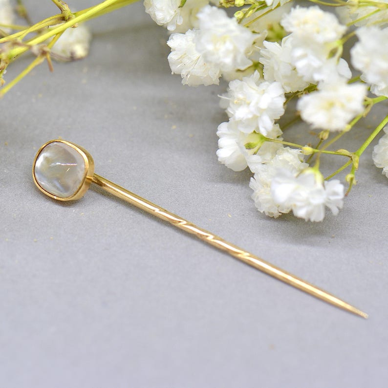 May include: A gold-colored hat pin with a square, iridescent stone at the top. The pin has a textured design on the shaft and a pointed end. White flowers are in the background.