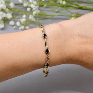 May include: A gold bracelet with dark blue teardrop-shaped gemstones and small white gemstones. The bracelet has a delicate, twisted design and is worn on a wrist. The background is a soft grey with white flowers.