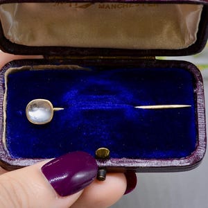May include: A vintage gold-tone stick pin with a square, translucent stone set in a gold frame. The pin is displayed in a velvet-lined box with a purple exterior. The box is open, revealing the pin against a deep blue background.