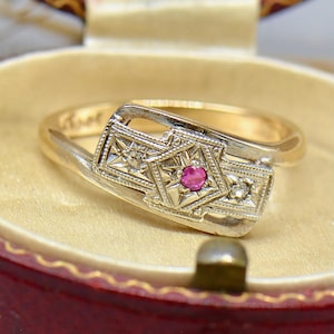 Puede incluir: Un anillo de oro vintage con un diseño único. El anillo presenta un engaste cuadrado central con una pequeña gema roja redonda, flanqueada por dos engastes cuadrados más pequeños con gemas claras. El anillo está en una caja de joyería roja.