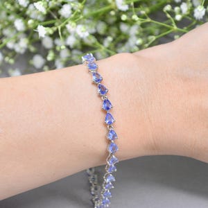 Può includere: Un braccialetto d'argento con una serie di pietre preziose blu chiaro a forma di pera. Il braccialetto è mostrato su un polso, con uno sfondo sfocato di fiori bianchi e una superficie grigia.